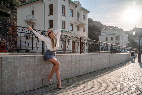 Outdoor photo of a romantic European blonde woman with long hair spending time outdoors exploring a European city. A graceful young lady enjoys the view of the embankment near the yacht