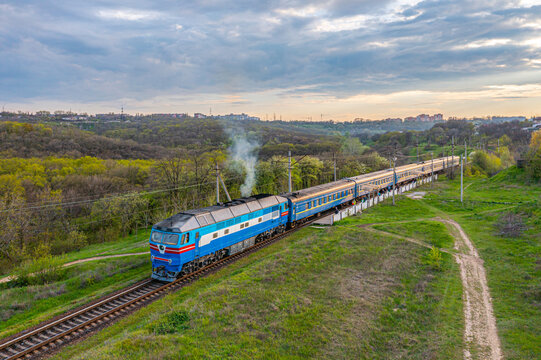 A freshly painted blue diesel locomotive with carriages of a passenger train departs from the platform of the stopping point. Aerial view. Spring photo.