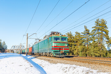 A powerful two-system green electric locomotive pulls a long freight train through a snow-covered pine forest in the sunset light. Winter photo.