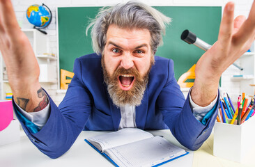 Angry Bearded teacher in classroom sits at desk. Back to school. Learning, education. Teachers day.