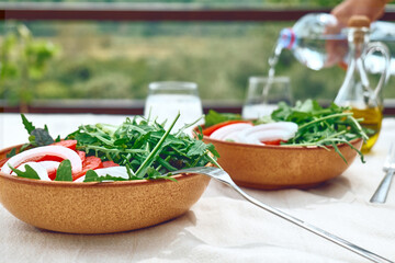 Healthy vegan salad with tomatoes, arugula, avocado, spinach red onion and olive oil. Summer refresh brunch or dinner. Defocused background where hand of man pouring water in the glass.