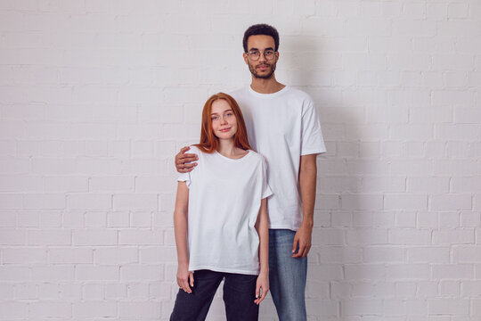 Beautiful international couple in white t-shirts stand on a white background.