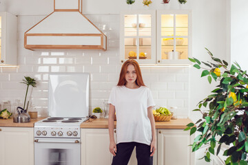 Young red-haired woman in a white t-shirt stands in the kitchen.