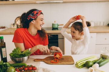 Mom and daughter cooking together, spending time at kitchen, young pretty dark-haired mother slicing bell peppers on board while her child fooling around, looking funny holding piece of red vegetable