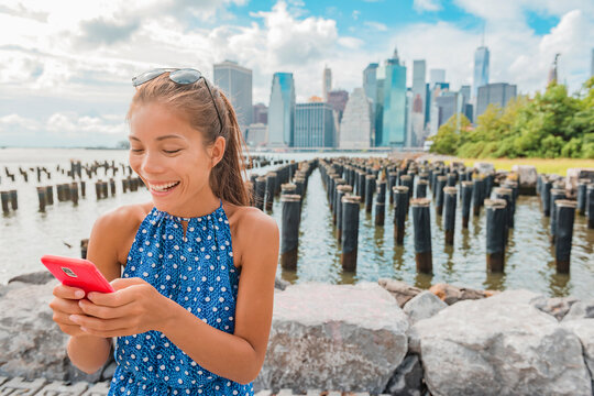 New York tourist woman using app on phone by Manhattan city skyline waterfront. People walking enjoying view of downtown from the Brooklyn Bridge park Pier 1 salt marsh