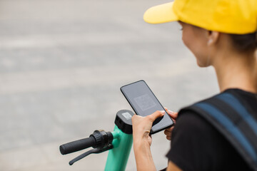 Unrecognizable woman in yellow cap and backpack using smartphone for renting modern electric...