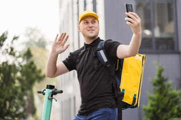 Man courier food delivery with yellow thermal backpack walks street with electric scooter uses...