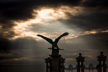 Sunlight breaking through the cloudy sky, shining on the eagle statue in front of the S&aacute;ndor Palace