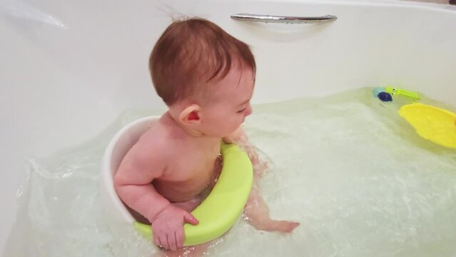 Happy toddler baby boy bathes in a white bath while sitting on a chair. Child playing with water in a large bathtub, kid is nine months old