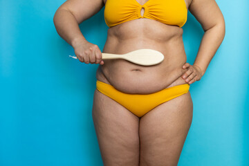 Unrecognizable fat plus-size overweight woman standing in yellow bra, underpants, swimsuit, brushing lymphatic drainage massage of stomach by wooden brush. Anticellulite treatment, homemade body care.