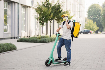 Side view of delivery man in face mask using smartphone and riding push scooter in the city. Young delivery man with thermal backpack using smartphone while riding scooter during covid pandemic.