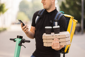 Close up of young delivery man calling by mobile phone to client outdoor. Courier delivering food and hot drinks outside. Guy using smartphone at city street. Courier with pizza and coffee.