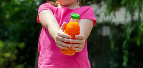 Anonymous young unrecognizable school age child, girl holding a glass bottle full of orange vegetable juice in hands, wide shot, object closeup. Giving, handing a drink, vitamins, healthy beverage