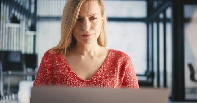 Caucasian mature woman in casual attire working on portable laptop while sitting at modern office. Beautiful blonde with focused facial expression looking on computer screen.