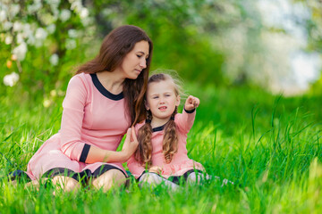 Fototapeta premium mother and daughter sit in the green grass against the backdrop of blooming apple trees