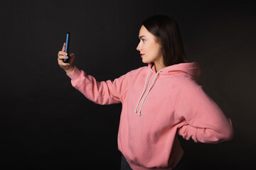 Cute young brunette in a pink hoodie uses a smartphone to take a selfie, portrait on a black background
