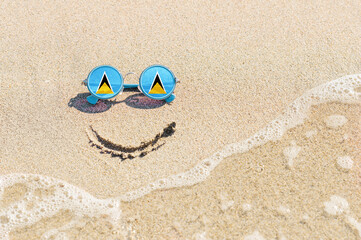 Fototapeta na wymiar A painted smile on the sand and sunglasses with the flag of the Saint Lucia. The concept of a positive and successful holiday in the resort of the Saint Lucia.