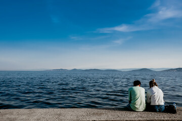 Fototapeta premium Back view of young couple sitting by the sea on beach. Desire, hope to go far away. Making holiday plan. Minimal Concept. Free area for text or ads