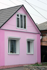 Pink facade of a one-story house with a triangular roof. Plastic windows in the house with metal blinds on top. The window in the attic.