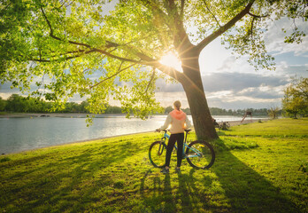 Woman riding a mountain bike near green trees and lake at sunset in spring. Colorful landscape with sporty girl, bicycle, beach, river, green grass, river in park. Summer. Sport and travel. Biking