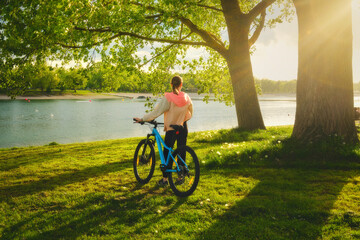 Woman riding a mountain bike near green trees and lake at sunset in spring. Colorful landscape with sporty girl, bicycle, beach, river, green grass, river in park. Summer. Sport and travel. Biking