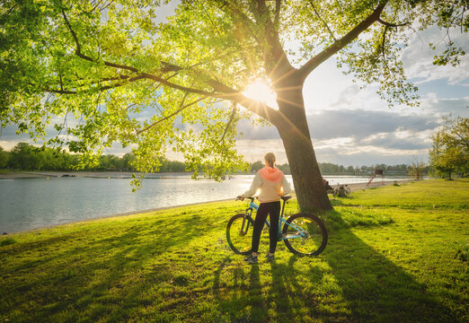 Woman Riding A Mountain Bike Near Green Trees And Lake At Sunset In Spring. Colorful Landscape With Sporty Girl, Bicycle, Beach, River, Green Grass, River In Park. Summer. Sport And Travel. Biking