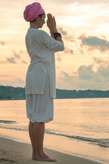 A woman in white clothes and a turban stands by the sea on a sandy beach at dawn on a summer morning and doing yoga exercises . Colorful sky on the background.