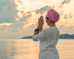 A woman in white clothes and a turban stands by the sea on a sandy beach at dawn on a summer morning and doing yoga exercises . Colorful sky on the background.