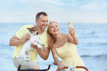 Mature couple with bicycles and dog taking selfie near sea on summer day