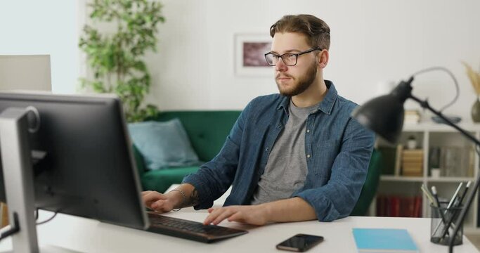 Professional programmer typing on pc keyboard while sitting at home office. Young bearded man working on new data code while sitting at desk.