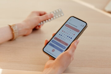 Close up of young woman calculating menstrual cycle using mobile app and holding birth control pills - obrazy, fototapety, plakaty