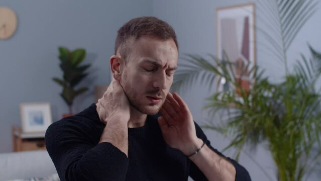 Close-up footage of Caucasian male employee sitting at table with laptop, holding neck while using computer. Remote work concept. Handsome young man suffering from neck pain at home. Indoors.