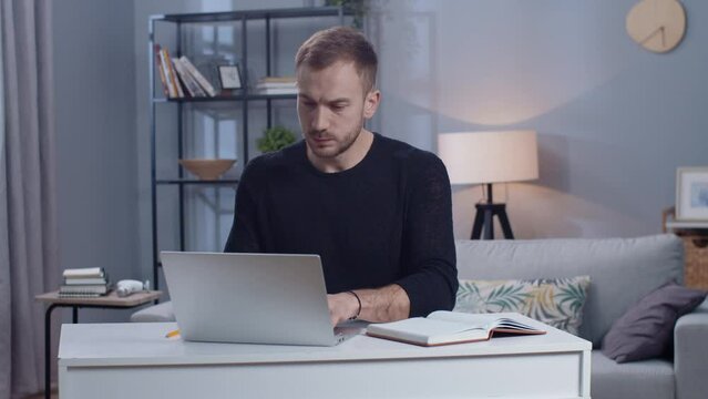 Portrait of focused young Caucasian man looking at screen of laptop, using computer at workplace at home. Serious businessman browsing. Guy suffering from neck pain. Casual outfit. Working day.