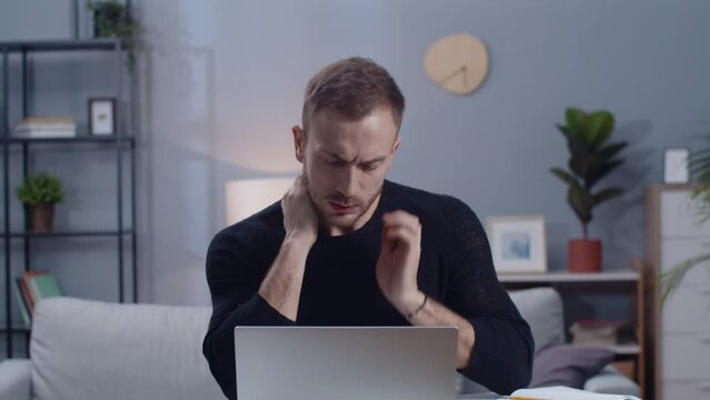 Close-up footage of tired young man holding neck while using computer, looking at notebook. Shot of good-looking guy in black sweatshirt sitting in comfortable living room. Ordinary working day.