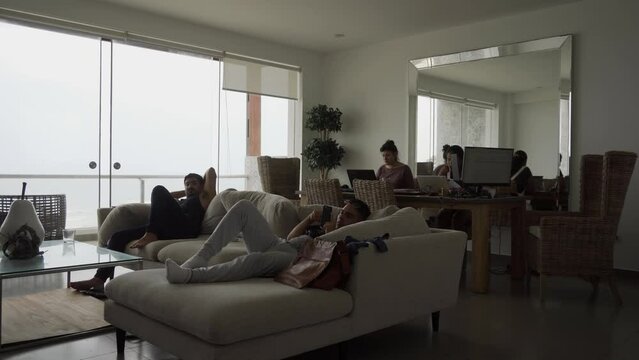 two friends resting in the armchair watching tv and their partners behind working with their laptops for homeoffice in a beach house with a background of the sea in the day in 4k