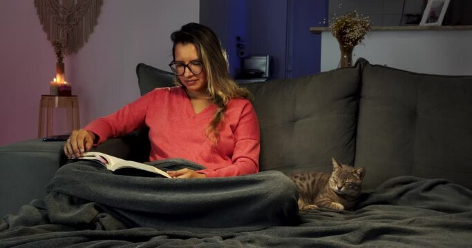 Mulher jovem lendo um livro sentada no sof&aacute; ao lado de um gato 