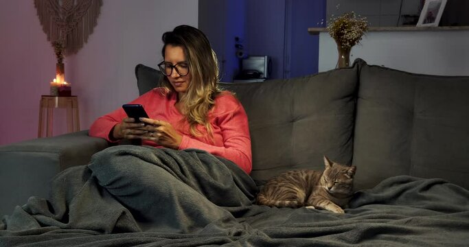 Mulher Jovem digitando no smartphone sentada no sof&aacute; ao lado de um gato