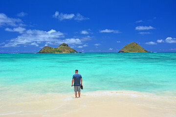 A lone tourist wading in turquoise waters while enjoying a lovely view of the Mokes aka Na Mokulua on a clear sunny day at Lanikai Beach on the windward side of Oahu, Hawaii.	. 