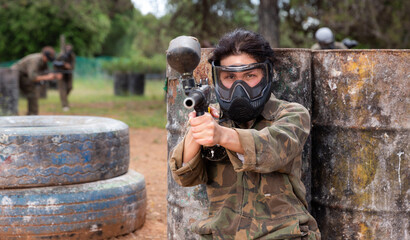 Portrait of girl paintball player in mask who is aiming in opponents on paintball field