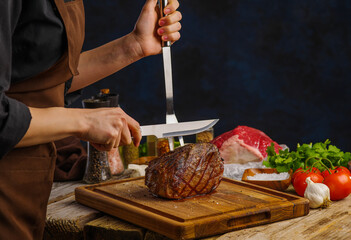 A professional chef cuts a cooked meat ham on a wooden cutting board on a blue background. Restaurant, hotel, home cooking. Recipes for restaurant and home cooking. Close-up.