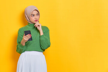 Pensive beautiful Asian woman in green sweater holding mobile phone looks seriously thinking about...