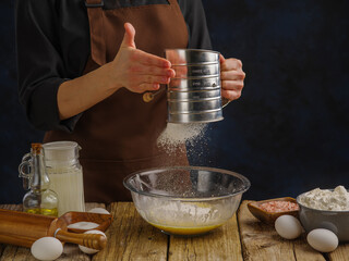 The chef is sifting flour through a sieve on a wooden kitchen table. Flour in frozen flight. Ingredients for making dough on a dark blue background. The concept is cooking from dough.