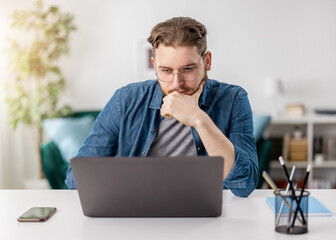 Pensive young caucasian man sitting at table and looking on computer screen. Male freelancer in eyewear browsing information in internet while working at home.