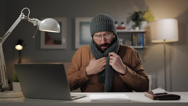 Cold at home. Low air temperature in living room, heating in apartment is turned off. Man in hat and scarf is cold indoors and works at computer