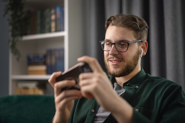 Obraz na płótnie Canvas Happy young man in wireless earphones sitting on couch and playing video games on smartphone. Relaxed caucasian guy using modern gadgets for taking fun at cozy apartment.