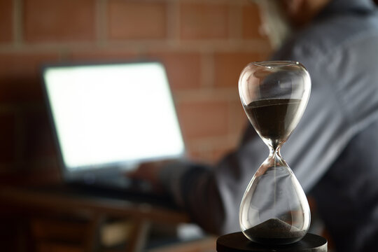 Hourglass and a man working on a laptop. Focus on close up sand glass. Concept for time management and countdown to deadline.