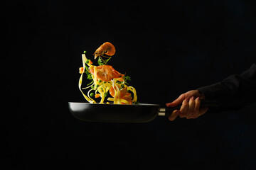 The process of cooking italian pasta with seafood and vegetables in a frying pan on a black background by a professional chef. Levitation. Recipes for healthy vegetarian food with seafood. - 502303618