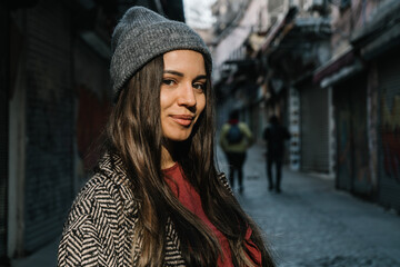 A student with dark long hair in a gray hat and a black and white coat. Walk through the streets of autumn Turkey. Portrait close-up on the chest against the background of the street. A happy girl