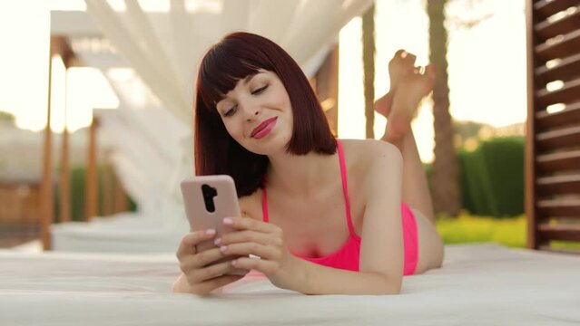 Video call from paradise. Joyful young lady in pink swimwear, lying in gazebo near the resort pool and using smartphone to make online video call to her friend or family.
