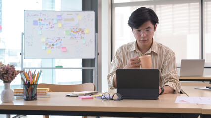 Focused young businessman drinking coffee and reading information on his computer tablet.